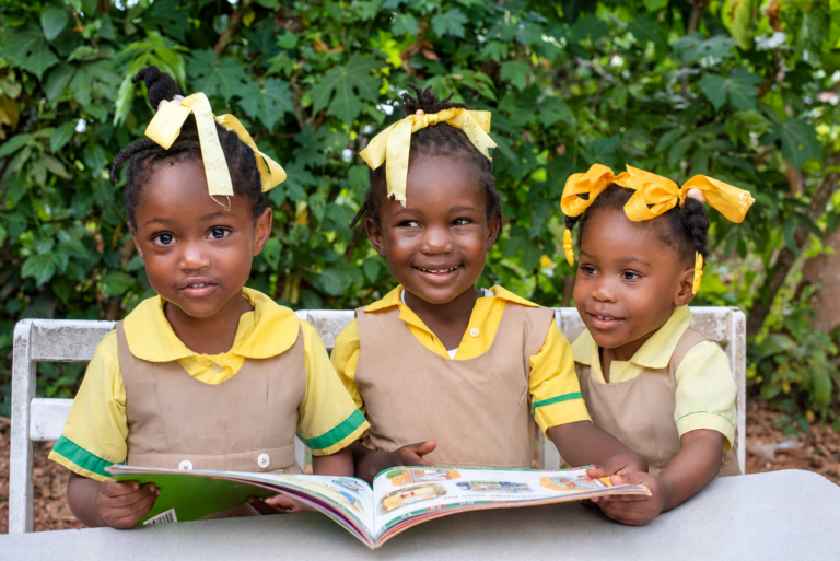 Three Academy children in yellow uniforms reading a book outdoors